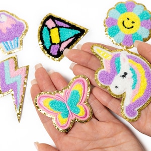 May include: A set of colorful iron-on patches featuring a unicorn, butterfly, lightning bolt, cupcake, flower, and a geometric shape. The patches are made of fuzzy fabric and have gold glitter trim.