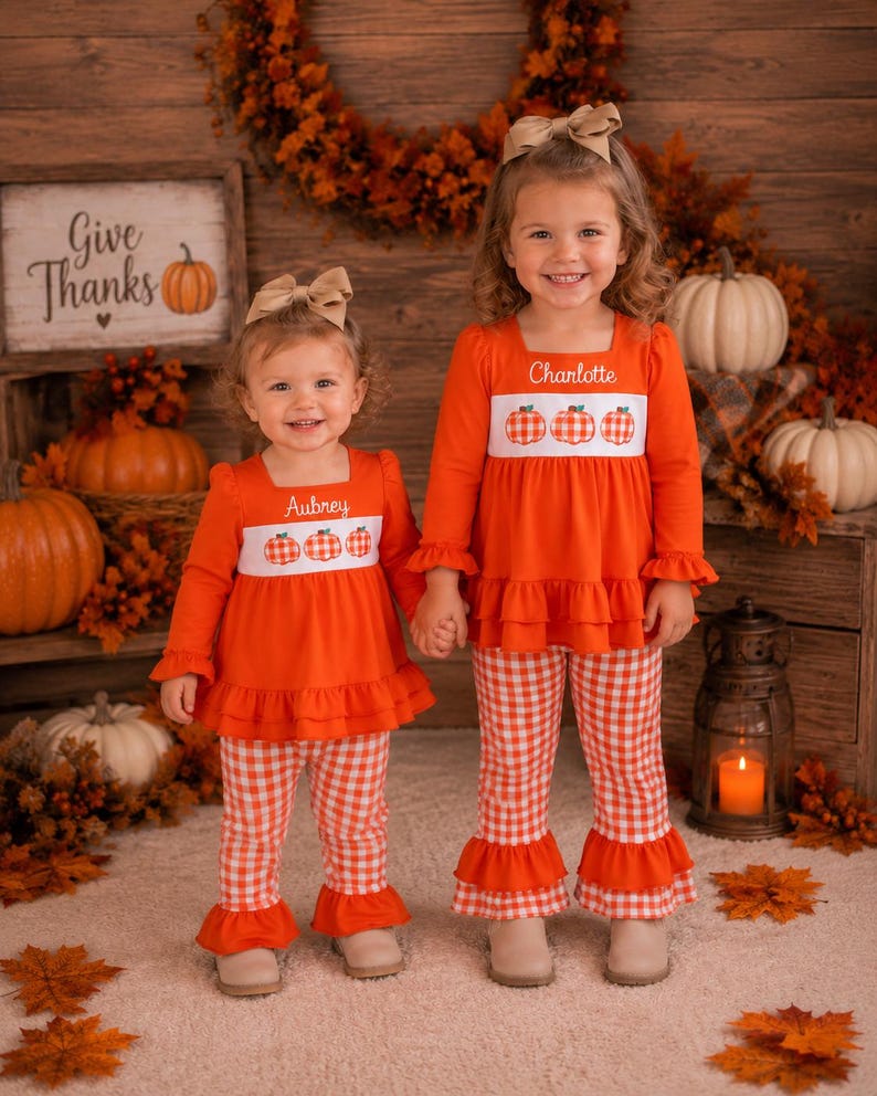 May include: Two matching orange long-sleeve shirts and orange and white gingham pants sets. Each shirt features a white chest panel with three embroidered pumpkins and a personalized name, Aubrey or Charlotte, in white script lettering.