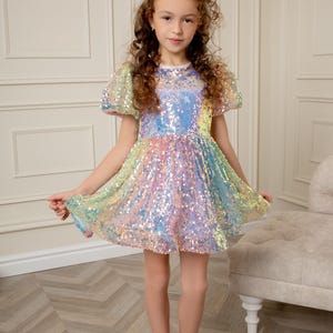Peut inclure: Une jeune fille portant une robe pailletée arc-en-ciel irisée à manches bouffantes courtes. Elle est debout devant un mur blanc et une chaise blanche.