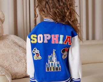 Puede incluir: Una chaqueta de béisbol azul y blanca con el nombre "SOPHIA" escrito en letras de colores en la espalda. La chaqueta tiene varios parches, incluyendo Mickey Mouse, Minnie Mouse y el castillo de Disney.