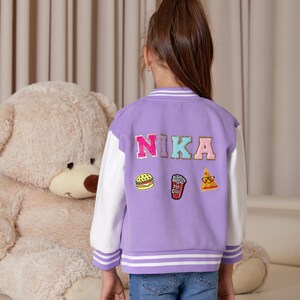 Puede incluir: Chaqueta de béisbol de color morado y blanco con el nombre "NIKA" bordado en la espalda. La chaqueta tiene tres parches en la espalda: una hamburguesa, palomitas de maíz y una pizza.
