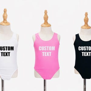 May include: Three children's swimsuits in white, pink, and black, each displayed on a mannequin. The swimsuits have a scoop neck and the text "CUSTOM TEXT" printed on the front in white. The mannequins have wooden tops.