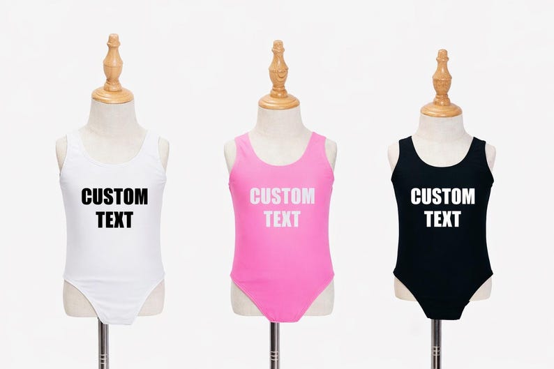 May include: Three children's swimsuits in white, pink, and black, each displayed on a mannequin. The swimsuits have a scoop neck and the text "CUSTOM TEXT" printed on the front in white. The mannequins have wooden tops.