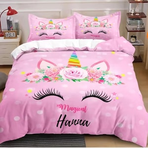May include: Pink unicorn-themed bedding set with a duvet cover and two pillowcases. The duvet features a large unicorn face with eyelashes, a rainbow horn, and the name "Hanna". The pillowcases have a smaller unicorn face design.