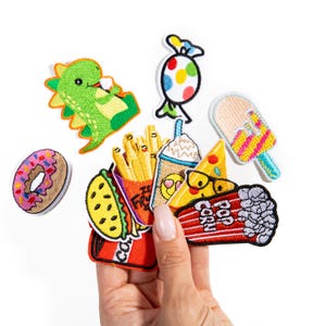 May include: A collection of colorful iron-on patches featuring various food and cartoon characters. The patches include a donut, a dinosaur, fries, a milkshake, a popsicle, a pizza slice, and a popcorn bucket.