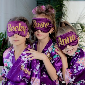 May include: Three young people wearing purple sleep masks with gold lettering that reads "Becky", "Rose", and "Anna". They are wearing matching purple robes with floral patterns. The image is a close-up shot.