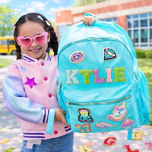 May include: A light blue backpack with the name "KYLIE" in glittery letters. The backpack has multiple patches, including a donut, a gem, a teddy bear, a butterfly, a lightning bolt, a unicorn, and a cupcake. The backpack has a front zipper pocket and a top handle.