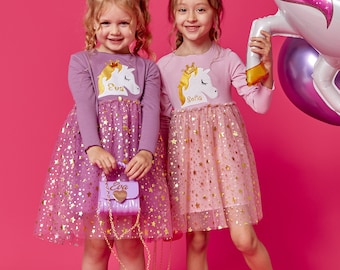 May include: Two girls wearing pink and purple dresses with unicorn and star designs. The girls are holding small purses with their names on them. The girl on the left is holding a purple purse with the name "Eva" on it. The girl on the right is holding a pink purse with the name "Sofia" on it.