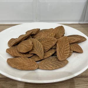May include: A white plate piled with brown, leaf-shaped items. Each item has a detailed vein pattern and a textured surface. The plate sits on a light-coloured wooden surface, with a white tiled background.