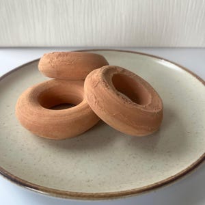 May include: Three terracotta-coloured, ring-shaped objects with a matte, porous texture. These circular items feature a central hole and a rounded, slightly uneven surface, arranged in a cluster on a speckled, off-white ceramic plate.