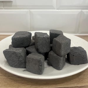 Puede incluir: Un plato blanco con una pila de briquetas de carbón vegetal negras. Las briquetas son cuadradas y tienen una textura rugosa.