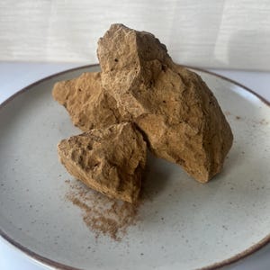 Könnte beinhalten: Drei unregelmäßige, hellbraune Tonstücke mit rauer, poröser Textur auf einem gesprenkelten, cremefarbenen Keramikteller. Eine kleine Menge feines, hellbraunes Pulver ist auf der Oberfläche des Tellers in der Nähe der Tonstücke verstreut.