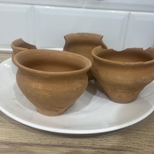 May include: Four small, unglazed terracotta pots of varying sizes and shapes sit on a white plate. The pots have a rustic, handmade appearance, with visible texture and imperfections. The pots are empty and ready for planting.