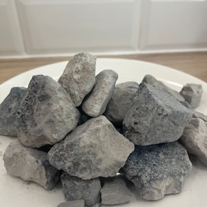 May include: A pile of gray, irregularly shaped rocks on a white plate. The rocks have a rough, textured surface and vary in size. The plate is round and sits on a light-colored surface, with a white tiled background.