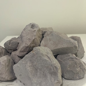 Puede incluir: Un montón de rocas grises de forma irregular, posiblemente arcilla, están dispuestas sobre un plato blanco. Las rocas tienen una superficie rugosa y texturizada y varían en tamaño. El fondo es una pared blanca lisa.