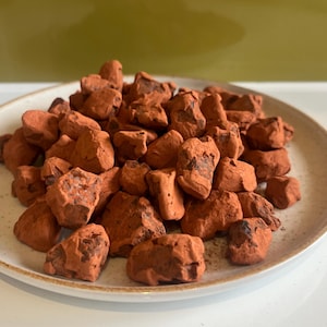 May include: A beige ceramic plate piled high with numerous, irregularly shaped, reddish-brown chocolate truffles. The truffles have a rough, textured appearance. The plate is set against a light background.