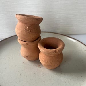 Puede incluir: Tres macetas pequeñas de terracota sin esmaltar con un acabado mate de color marrón tierra. Las macetas presentan un cuerpo redondeado, una abertura ancha con un borde grueso y una superficie texturizada. Dos están apiladas y una al lado.
