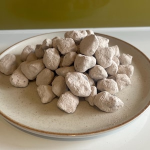 May include: A beige ceramic plate filled with a pile of light-colored, irregularly shaped rocks. The rocks have a slightly textured surface and are clustered together. The plate has a simple design with a brown rim, and the background is a solid green.