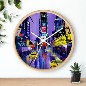 Puede incluir: Un reloj redondo con fondo morado y una pintura de Times Square en la ciudad de Nueva York. La esfera del reloj tiene agujas blancas y números negros. La pintura incluye taxis amarillos, farolas y edificios con letreros de espectáculos de Broadway como "Aladdin" y "El Rey León".