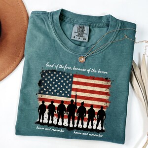 May include: A teal blue t-shirt with a distressed American flag design and a line of soldiers in silhouette. The text "land of the free, because of the brave" and "honor and remember" are printed on the shirt.