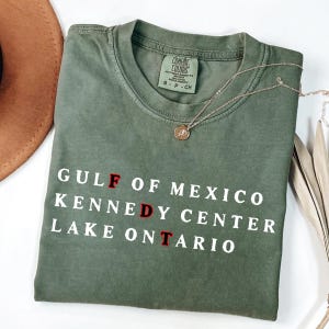 May include: An olive green short-sleeve t-shirt with white text that reads GULF OF MEXICO, KENNEDY CENTER, and LAKE ONTARIO. The letters F, D, and T are highlighted in red. The shirt features a Comfort Colors brand label at the inner neckline.