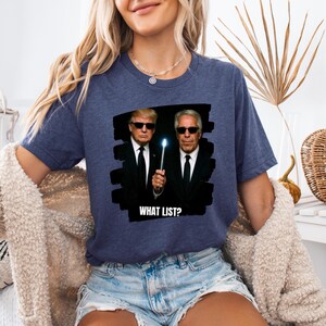 K&ouml;nnte beinhalten: Marineblaues T-Shirt mit einer Grafik von zwei M&auml;nnern in schwarzen Anz&uuml;gen und Sonnenbrillen, die einen leuchtenden Zauberstab halten. Der Text "WHAT LIST?" steht darunter. Das T-Shirt ist ein l&auml;ssiger Stil.