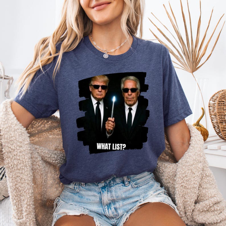 K&ouml;nnte beinhalten: Marineblaues T-Shirt mit einer Grafik von zwei M&auml;nnern in schwarzen Anz&uuml;gen und Sonnenbrillen, die einen leuchtenden Zauberstab halten. Der Text "WHAT LIST?" steht darunter. Das T-Shirt ist ein l&auml;ssiger Stil.