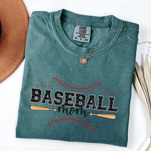 May include: A teal blue t-shirt with the text "Baseball Mom" in black with red stitching around the text. Two wooden baseball bats are crossed behind the text.