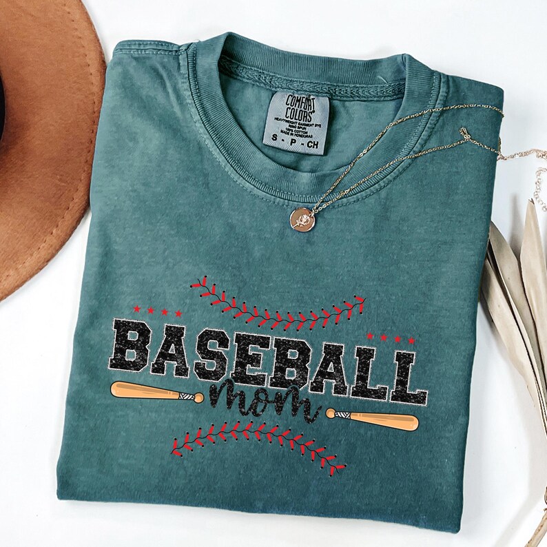 May include: A teal blue t-shirt with the text "Baseball Mom" in black with red stitching around the text. Two wooden baseball bats are crossed behind the text.