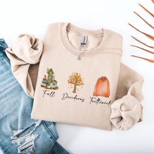 Może przedstawiać: Beżowa bluza z ilustracjami i tekstem w jesiennym klimacie. Projekt zawiera domek dla ptaków, drzewo i golf, z napisami "Fall", "Deciduous" i "Turtleneck" poniżej. Srebrny naszyjnik jest zarzucony na bluzę.