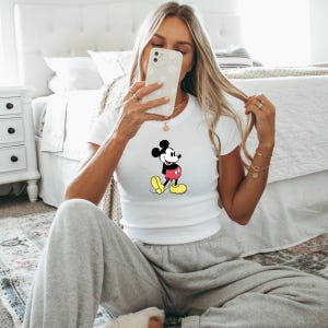 May include: White t-shirt featuring a classic Mickey Mouse graphic. The cartoon character is depicted in black, red, yellow, and white. The shirt is short-sleeved and appears to be made of a soft material.