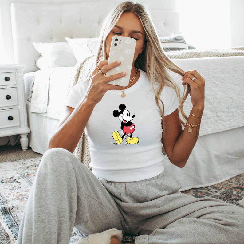 May include: White t-shirt featuring a classic Mickey Mouse graphic. The cartoon character is depicted in black, red, yellow, and white. The shirt is short-sleeved and appears to be made of a soft material.