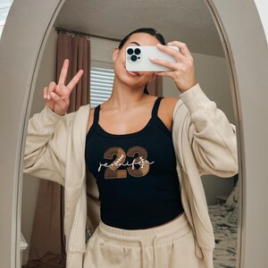 May include: A black ribbed tank top featuring the number 23 in a brown football-themed pattern with white stitching details, layered under a beige zip-up hoodie. The name Jennifer is written in white cursive script across the center of the number 23.