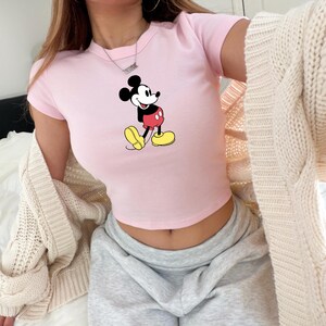 May include: A light pink crop top featuring a graphic of Mickey Mouse in his classic pose. The cartoon character is depicted in black, red, yellow, and white. The top has short sleeves and a crew neck.