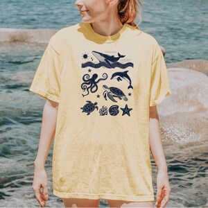 May include: Yellow t-shirt featuring a navy blue graphic of various sea creatures, including a whale, dolphin, octopus, and sea turtles. The design also includes a sun, stars, and other marine elements. The shirt is made of a soft material.