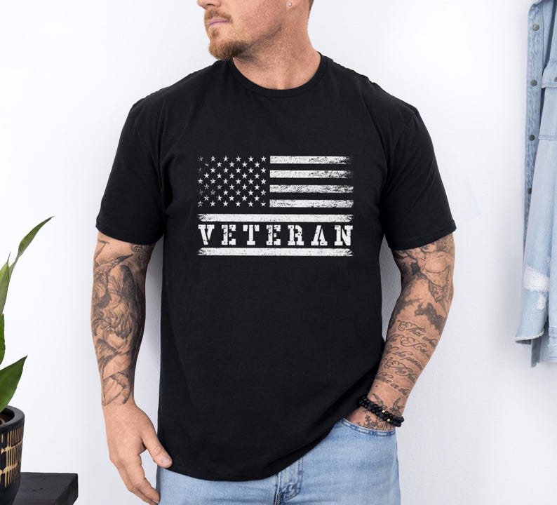 以下が含まれることがあります： アメリカの旗のデザインがくたびれた黒のTシャツで、白い文字で「VETERAN」と書かれている。