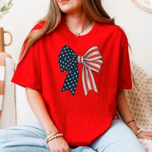 Peut inclure: T-shirt rouge avec un grand n&oelig;ud imprim&eacute; sur le devant. Le n&oelig;ud est fait de tissu rouge et bleu avec un motif d'&eacute;toiles blanches.