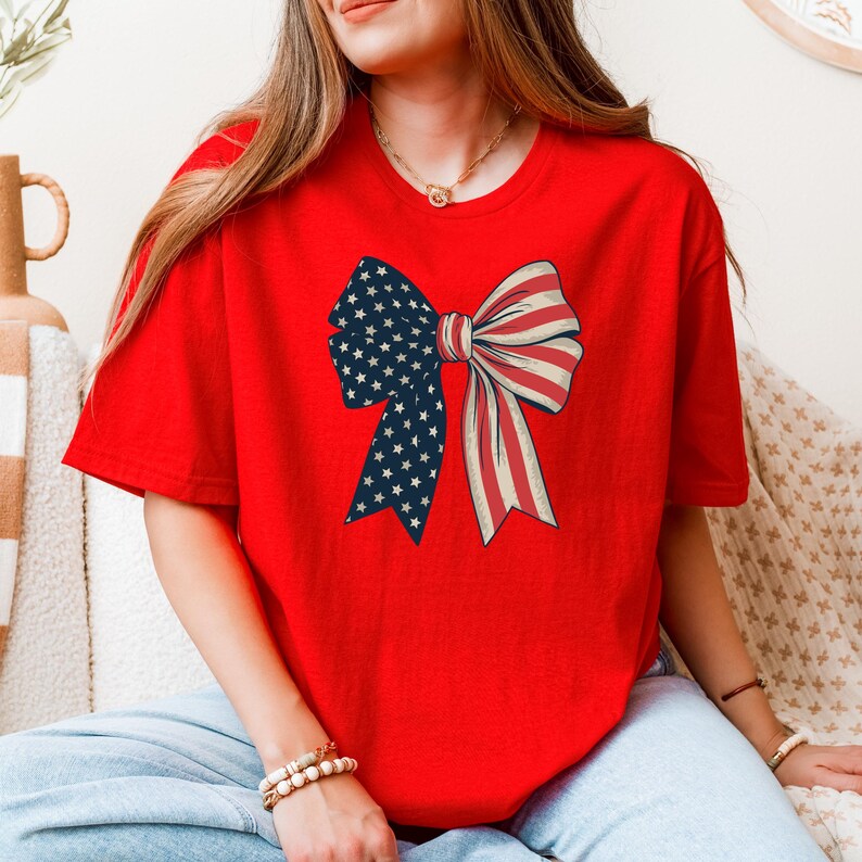 Peut inclure: T-shirt rouge avec un grand n&oelig;ud imprim&eacute; sur le devant. Le n&oelig;ud est fait de tissu rouge et bleu avec un motif d'&eacute;toiles blanches.
