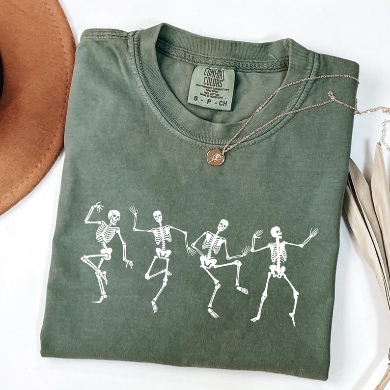 May include: An olive green Comfort Colors t-shirt with a white graphic of dancing skeletons. A gold necklace with a pendant is draped across the shirt. The shirt is folded, with a brown hat visible in the background.