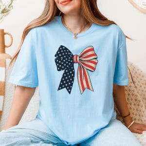 Peut inclure: Un t-shirt bleu clair avec un n&oelig;ud rouge, blanc et bleu. Le n&oelig;ud est con&ccedil;u pour ressembler &agrave; un drapeau am&eacute;ricain.
