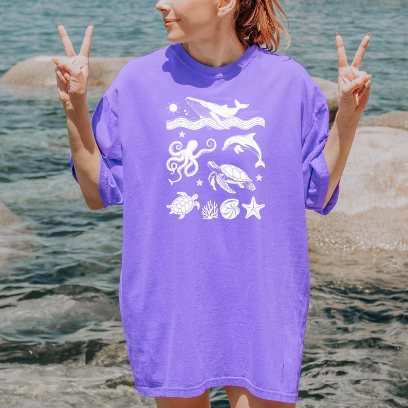 May include: A purple t-shirt featuring a white graphic of various sea creatures, including a whale, octopus, dolphin, and sea turtle. The design also includes a starfish, coral, and a sun. The shirt is a solid color and has a relaxed fit.