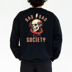 May include: Black sweatshirt with a graphic print of a skull with a snake wrapped around it. The text "Rad Dad Society" is printed in brown letters on the back of the sweatshirt.