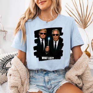 K&ouml;nnte beinhalten: Hellblaues T-Shirt mit einer Grafik von zwei M&auml;nnern in schwarzen Anz&uuml;gen und Sonnenbrillen, einer h&auml;lt einen leuchtenden Zauberstab. Der Text "WHAT LIST?" steht darunter. Das Shirt ist aus weichem Material.