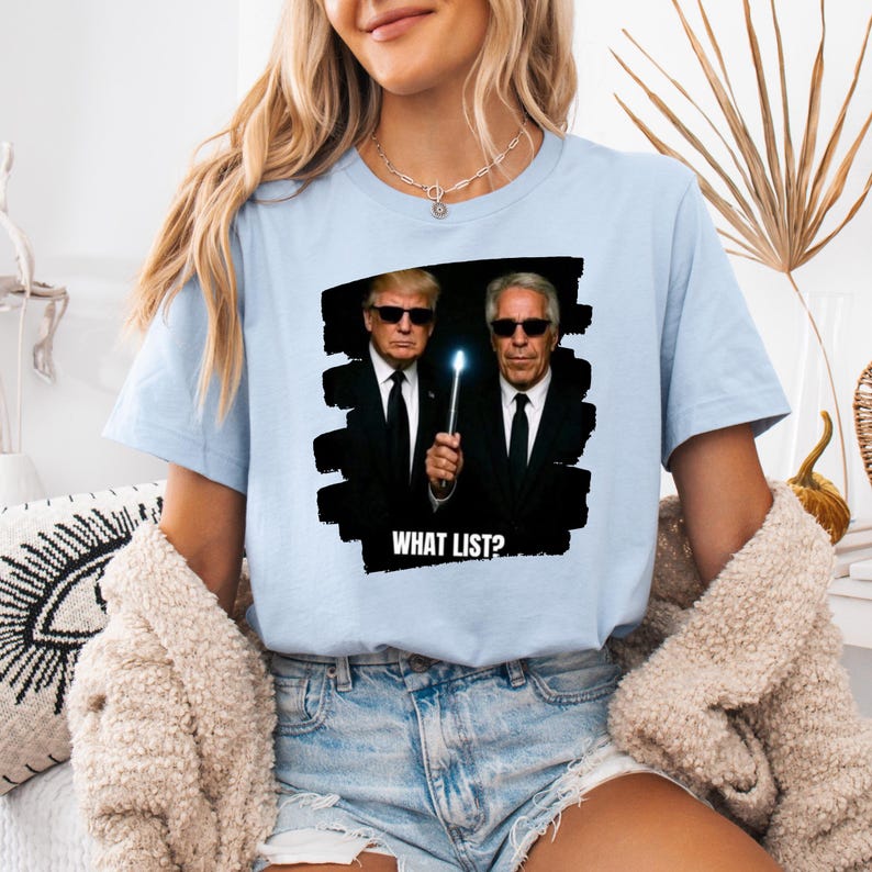 K&ouml;nnte beinhalten: Hellblaues T-Shirt mit einer Grafik von zwei M&auml;nnern in schwarzen Anz&uuml;gen und Sonnenbrillen, einer h&auml;lt einen leuchtenden Zauberstab. Der Text "WHAT LIST?" steht darunter. Das Shirt ist aus weichem Material.