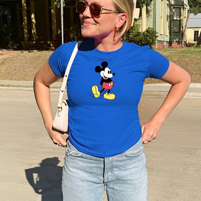 May include: A royal blue t-shirt featuring a graphic of Mickey Mouse. The cartoon character is depicted in his classic outfit with red shorts, yellow shoes, and white gloves. The shirt is paired with light-wash denim jeans.