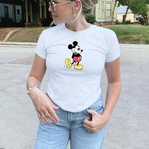 May include: White t-shirt featuring a classic Mickey Mouse graphic. The cartoon character is depicted in his iconic outfit: black shorts, yellow shoes, and red pants. The shirt is a crew neck style.