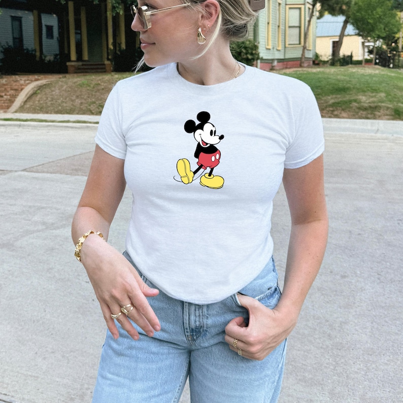 May include: White t-shirt featuring a classic Mickey Mouse graphic. The cartoon character is depicted in his iconic outfit: black shorts, yellow shoes, and red pants. The shirt is a crew neck style.