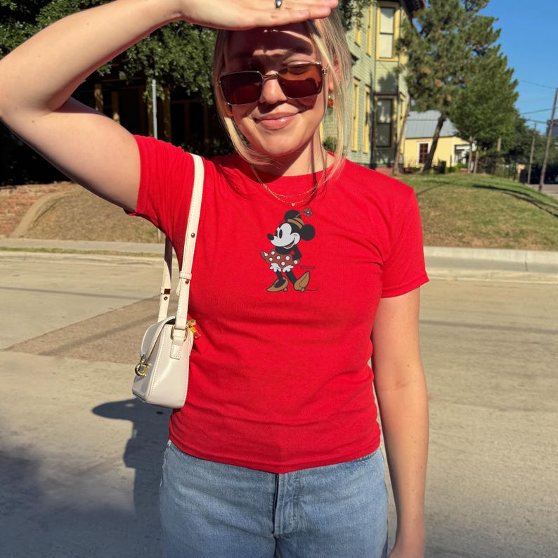 May include: A red t-shirt featuring a Minnie Mouse graphic. The graphic shows Minnie Mouse in a polka dot dress and bow. The shirt is paired with blue jeans and a white purse.