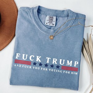 May include: A blue Comfort Colors t-shirt with the text "FUCK TRUMP" in white, along with the phrase "AND FUCK YOU FOR VOTING FOR HIM". The text is above a red and blue star design. A gold necklace is draped across the shirt.