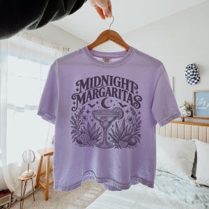May include: A light purple short-sleeve t-shirt featuring a dark purple graphic print on the front. The design displays a margarita glass, agave plants, lime slices, stars, bats, and a crescent moon with the text Midnight Margaritas.